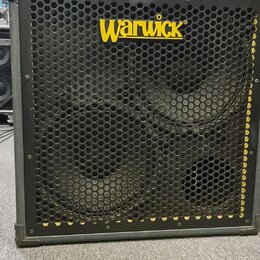 Černý basový reproduktor v šedém rámu s kovovou ochrannou mřížkou. V horní části je žluté logo Warwick. Za mřížkou jsou vidět dva velké reproduktory…