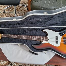 V otevřeném černém kufru s vnitřním čalouněním leží elektrická basová kytara. Nástroj má tělo v barvě sunburst, bílý pickguard a hnědý hmatník. Na hl…