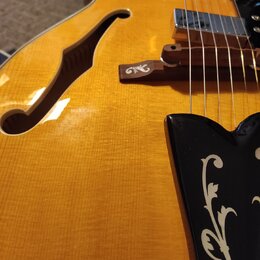 Detailní pohled na tělo žluté polodupaté elektrické kytary s f-otvorem. Na dřevěném kobylníku je viditelné logo znacky Epiphone. Kytara má dva chromo…