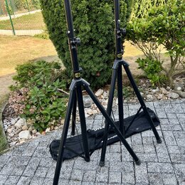 Dvě černé kovové stativy na hudební nástroje s teleskopickými horními trubicemi. Na obou stativech je viditelné malé žluté kulaté logo s nápisem HARL…