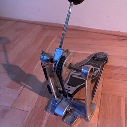 Kovový basový pedál s černým šlapadlem a bílo-černým beaterem. Na bočním kole jsou vidět nápisy "SUPERGLIDE" a "CHAIN DRIVE". Povrch kovových částí v…