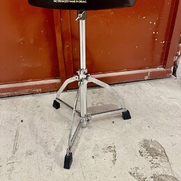 Černý bubenířský židle s kovovým chromovaným stojanem a třínožkou. Na sedáku je bílý nápis TAMA a pod ním text THE STRONGEST NAME IN DRUMS. Povrch se…