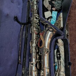 Stříbrný saxofon v otevřeném kufru s fialovým vnitřkem. K nástroji patří munzpun s mouthpiecem a samostatný hnědý řemínek. V kufru jsou rovněž umístě…