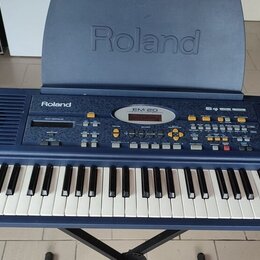 Modrý elektronický klavír umístěný na černém stojanu. Na horním panelu je nápis Roland a označení modelu EM-20. Nástroj má bílé a černé klávesy, řadu…
