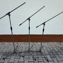 Tři šedé mikrofonní stojany s rameny. Stojany mají trojnožku a teleskopické části s černými utahovacími knoflíky. Povrch je matný, bez viditelných zn…