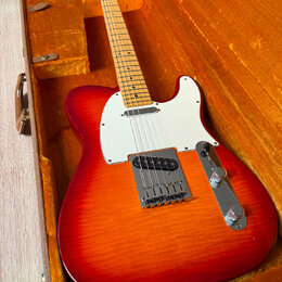 Elektrická kytara s červeno-oranžovým gradientem (sunburst) v hnědém kufru s oranžovou výplní. Nástroj má bílý pickguard, javorový hmatník a chromova…