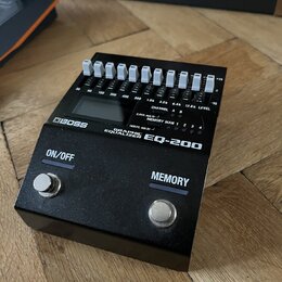 Černý grafický ekvalizér s bílými posuvníky a dvěma stomečními spínači. Na přístroji jsou nápisy BOSS, GRAPHIC EQUALIZER EQ-200, ON/OFF a MEMORY. Nad…