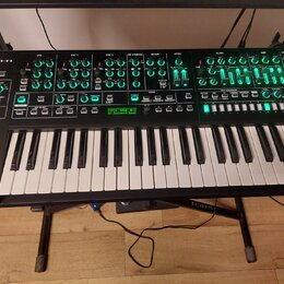 Černý klávesový syntetizátor s bílými a černými klávesami, umístěný na černém stojanu. Na levé straně horního panelu je bílý nápis Roland SYSTEM-8 PL…