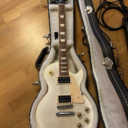Kytara s bílým trupem a hnědým hmatníkem v černé kostelní kytarové skříni. Na hlavě je viditelné logo Gibson. Nástroj má dva humbuckery, čtyři otočné…