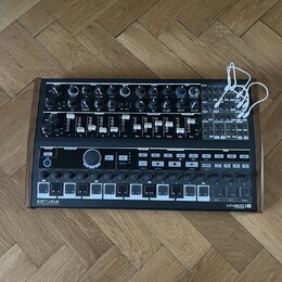 Černý analogový syntetizátor v dřevěném rámu s množstvím potenciometrů, posuvníků a tlačítek. Na spodním panelu jsou vlevo nápis ARTURIA a vpravo MIN…
