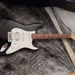 Černá elektrická kytara s bílým pickguardem a hnědým hmatníkem. Na hlavě nástroje je viditelné logo Fender. Kytara má tři snímače, pětistupňový přepí…