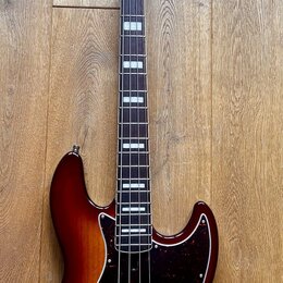 Elektrická basová kytara s trupem v červenohnědém sunburst provedení a hnědým hmatníkem s bílými polohovými značkami. Nástroj má tmavě červený pickgu…
