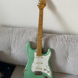 Světle zelená elektrická kytara s bílým pickguardem a javorovým hmatníkem. Na hlavě je viditelné logo Fender. Lak na těle vykazuje výrazné opotřebení…
