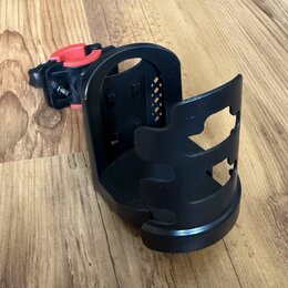 Černý plastový držák na příslušenství s červeným utahovacím mechanismem. Předmět je umístěn na dřevěné podlaze. Na povrchu nejsou viditelné žádné zna…
