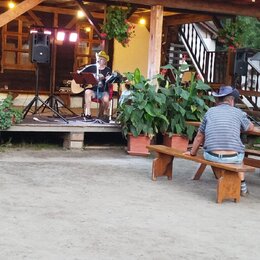 Fotografie zachicuje muzikanta na dřevěném pódiu, který hraje na akustickou kytaru světlé barvy. Vedle něj je umístěn mikrofon na stativu a černý rep…