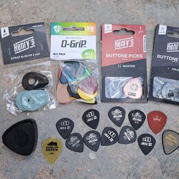 Fotografie zobrazuje sadu kytarového příslušenství. Jsou zde čtyři originální balení: „Henry's STRAP BLOCKS 2 mm“, „JANICKER PICKS D-Grip™ MIX PACK N…