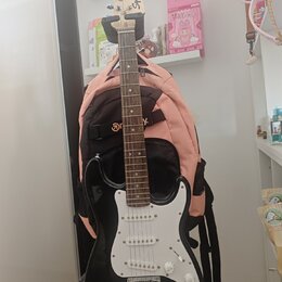 Černá elektrická kytara s bílým pickguardem a světle hnědým hmatníkem. Na hlavě nástroje je viditelné logo Squier. Kytara stojí na stojanu před růžov…