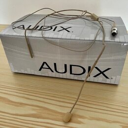 Na bílé krabici s nápisem AUDIX leží béžový hlavní mikrofon s tenkým ramínkem a konektorem. Na krabici je logo AUDIX zopakováno dvakrát. Povrch krabi…