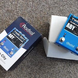 Modrý kovový pedál s nápisy TWIN-CITY CLASS-A AMP SWITCHER a ABY. Na spodní části je logo Radial engineering a přepínač označený jako TOGGLE. Vedle p…