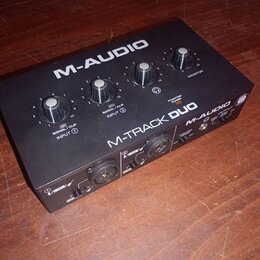 Černý audio rozhraní s nápisy M-AUDIO a M-TRACK DUO. Na horní straně jsou čtyři ovládací knofíky a kontrolky s popisky SIGNAL / CLIP INPUT 1, SIGNAL…