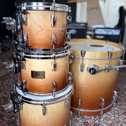 Sada bubnů v oranžovém přechodovém odstínu s viditelnou strukturou dřeva. Na prostředním tomu je kovový štítek s nápisem Pearl Masters Custom. Horní…