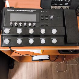 Černý kytarový procesor s nápisem BOSS GT-1000 GUITAR EFFECTS PROCESSOR a logem A.I.R.D. Zařízení má displej, pět malých tlačítek s čísly 1 až 5 a de…