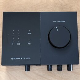 Černý audio rozhraní s matným povrchem. Na horní části jsou nápisy OUT 1/2 VOLUME, USB, 48 V a označení vstupů IN 1 a IN 2 u sloupců s kontrolkami. V…