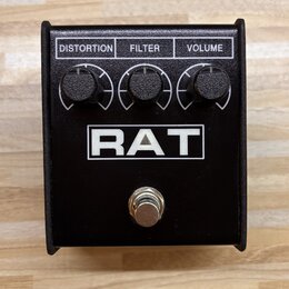 Černý efekt pedál s nápisem RAT v bílém obdélníku. V horní části jsou tři černé otočné ovladače s popisky DISTORTION, FILTER a VOLUME. Ve spodní část…