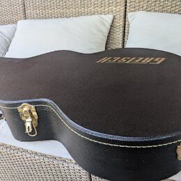 Černý tvrdý kufřík na kytaru s texturovaným povrchem a bílým prošíváním po okrajích. Na horní části je zlatý nápis GRETSCH. Kufřík má zlaté kovové zá…