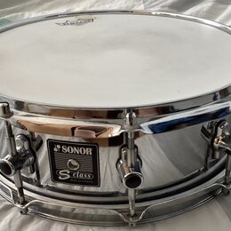 Stříbrný chromovaný snare drum s bílými koženem. Na korpusu je čitelný nápis SONOR a pod ním štítek s textem S class MADE IN GERMANY. Nástroj vykazuj…
