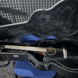 Černá akustická kytara s cutawayem v černém tvrdém kufru s polstrováním. Na hlavě hmatníku je viditelné logo Taylor. Na vnitřní straně kufru je nalep…