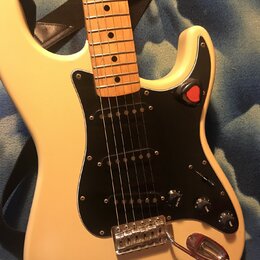 Elektrická kytara s krémovým trupem, černým pickguardem a hmatníkem ze světlého dřeva. Nástroj má tři snímače typu single-coil, jeden přepínač a tři…