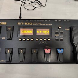 Černý kytarový procesor s nápisem BOSS GT-100 COSM AMP EFFECTS PROCESSOR. Zařízení má displej s oranžovým textem BOSS GT-100 Ver. 2, několik ovládací…