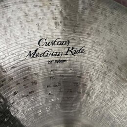 Detailní pohled na kovový činel s viditelným kovaným povrchem. Uprostřed je černý nápis „Custom Medium Ride“ a pod ním označení „22"/56cm“. Povrch vy…