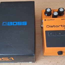 Kytarový efekt v oranžovém kovovém provedení s černým pedálem. Na zařízení jsou nápisy Distortion, DS-1 a logo BOSS. Vedle efektu je černá krabice s…