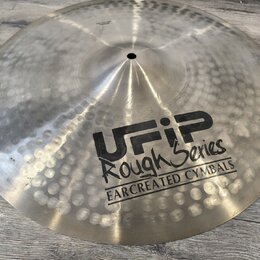 Kovový činel s kovaným povrchem a viditelnými důlky. Na povrchu je černý nápis UFIP Rough Series EARCREATED CYMBALS. V horní části je malý, nečitelný…