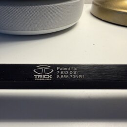 Detailní záběr na černý kovový povrch s bílými nápisy. Je zde vidět logo a text TRICK DRUMS USA. Vedle nich jsou uvedeny nápisy Patent No. 7,633,000…