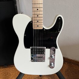 Bílá elektrická kytara typu Telecaster s černým plechtrátem a hnedým javorovým hmatníkem. Nástroj má chromovaný most s integrovaným snímačem a ovláda…