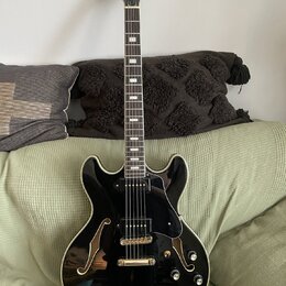 Černá polodublová elektrická kytara s lesklým povrchem a f-otvory. Na hlavě hmatníku je zlatý nápis "Joe Pass". Nástroj má zlaté ladičky, černý pickg…