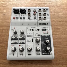 Bílá mixovací konzole značky YAMAHA, model AG06 MIXING CONSOLE. Zařízení obsahuje různé vstupy a výstupy, včetně konektorů XLR, jack 6,35 mm a USB. N…