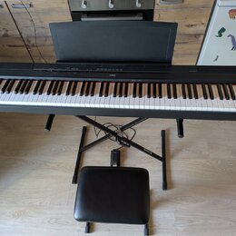 Černý digitální piano s klávesnicí, umístěné na černém X-stojanu s přiloženou černé polstrovanou židličkou. Na horní liště nástroje je uprostřed bílý…