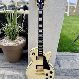 Elektrická kytara s krémovým povrchem korpusu a hmatníkem z tmavého dřeva s bílými blokovými vložkami. Na hlavě je zlatý nápis „Gibson“ a modrý ornam…
