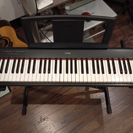 Černý digitální klavír značky YAMAHA umístěný na černém stojanu. Nástroj má bílé a černé klávesy s červeným pruhem nad nimi. V horní části je umístěn…