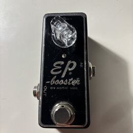 Černý kytarový efekt pedál v kovovém šasi. Na povrchu je bílý nápis „EP -booster BY xotic usa.“ a označení vstupů „IN“ a „OUT“. V horní části je průh…