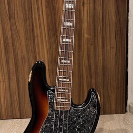 Elektrická basová kytara s trupem v barevnosti sunburst a černým pickguardem s texturou. Na hlavě hmatníku je nápis Fender Jazz Bass. Nástroj vykazuj…