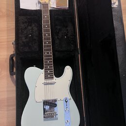Světle modrá elektrická kytara typu Telecaster v černém tvrdém kufru. Na hlavě je viditelné logo Fender. Kytara má bílý pickguard, chromovaný most a…