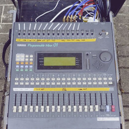 Šedý míchací pult s nápisem YAMAHA Programmable Mixer 01. Horní část obsahuje řadu přepínačů s žlutými papírovými štítky, uprostřed je umístěn LCD di…