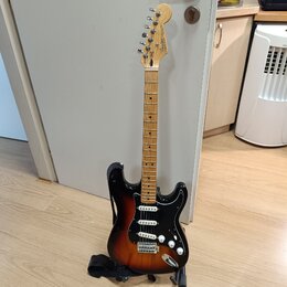 Elektrická kytara s trupem v barevnosti sunburst a černým pickguardem. Na hlavě hmatníku je viditelné logo Fender. Nástroj má světle hnědý krk a hmat…