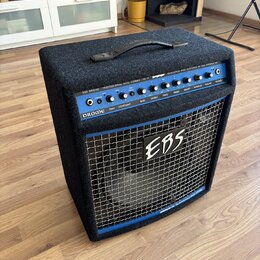 Černý basový kombo zesilovač s modrým ovládacím panelem a kovovou mřížkou. Na horní části je nápis EBS DROME PROFESSIONAL BASS COMBO 750 W a DEVELOPE…