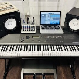 Digitální piano šedé barvy s nápisem ROLAND DIGITAL PIANO RD-1000, umístěné na černém stojanu s pedály. Na nástroji jsou umístěny dva černé studiové…